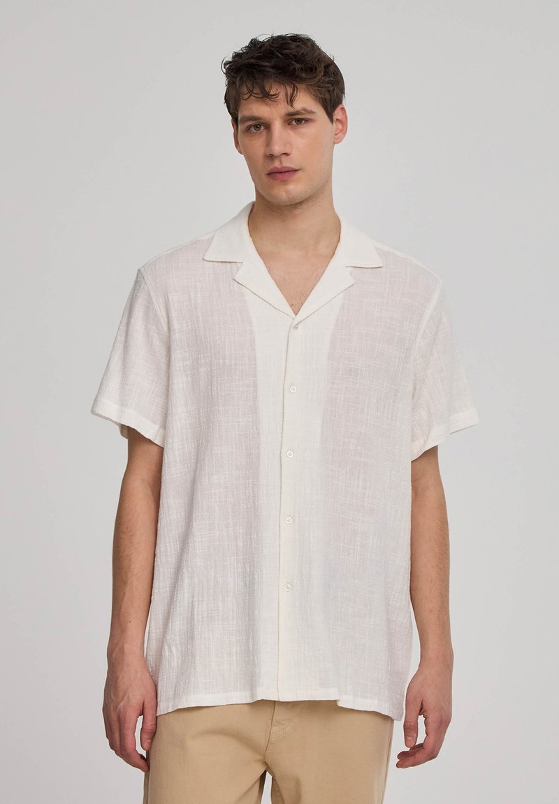 Camicia bianca di lino a maniche corte con colletto a punta, bottoni frontali e un motivo testurizzato. Indossata con pantaloni beige.