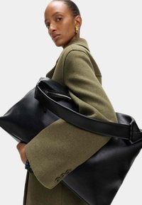 Zwarte leren tote bag met een ritssluiting en brede schouderband, vastgehouden tegen een groene jas met een gestructureerde vorm en zachte textuur.