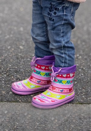 Kind, das eine blaue Jeans und bunte pinke Gummistiefel mit Herzmustern trägt, steht auf einer Asphaltfläche.