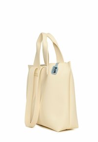 Sac fourre-tout en simili cuir beige avec deux poignées courtes et une sangle longue amovible. Texture lisse, design minimaliste et un détail de tag bleu.