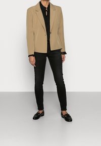 Béžový blazer s klopami, zapínání na jeden knoflík, nošený přes černou blůzu s volány, v kombinaci s černými skinny džínami a černými mokasíny.