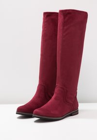 Bottes bordeaux montantes jusqu'au genou en matière douce et veloutée. Elles présentent un talon plat, un bout arrondi et des détails de couture sur la tige.