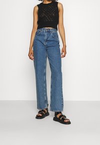 Schwarzes gestricktes Crop-Top mit Spitze-Design, kombiniert mit hochgeschnittenen blauen Denim weitgeschnittenen Jeans. Schwarze Sandalen mit Riemen.