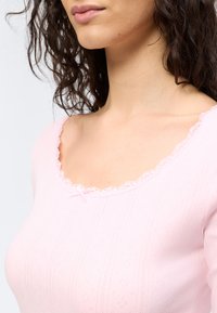 Γυναίκα με σγουρά σκουρόχρωμα μαλλιά που φοράει ανοιχτό ροζ μπλουζάκι με κυματιστή λαιμόκοψη και μικρή λεπτομέρεια φιόγκου.