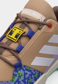 Tanfarvet sneaker med brune snørebånd, blå og grøn pixeleret mønster, hvide striber og gul Minecraft-inspireret snørebåndsvedhæng.