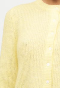 Μαλακό κίτρινο πλεκτό ζακέτα με λευκά κουμπιά, ριμπ λαιμόκοψη και μακριά μανίκια, απεικονίζεται στο πάνω μέρος του κορμού και της περιοχής του λαιμού.