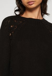 Suéter negro de punto con cuello redondo y detalles calados en los hombros. La textura es suelta y texturizada, lo que proporciona transpirabilidad.