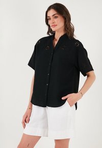 Black short-sleeve shirt with button-down front, featuring floral embroidery on shoulders and textured fabric. Paired with white shorts.