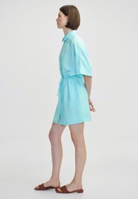 Robe bleu clair à manches courtes, avec un cordon de serrage à la taille et un ourlet légèrement évasé. Associée à des sandales brunes à lanières.