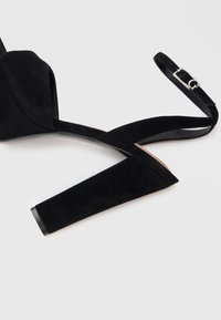 Zwarte suède hoge sandalen met blokhak en verstelbare enkelriem met een zilveren gesp op een witte achtergrond.