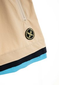 Chaqueta beige con cremallera vertical, parche circular con logo negro y amarillo, y bordes de tela superpuestos en negro y azul.