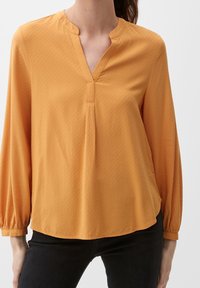 Blouse jaune moutarde avec col en V, détail plissé et manches longues. Tissu doux avec une texture subtile. Portée avec un pantalon noir.