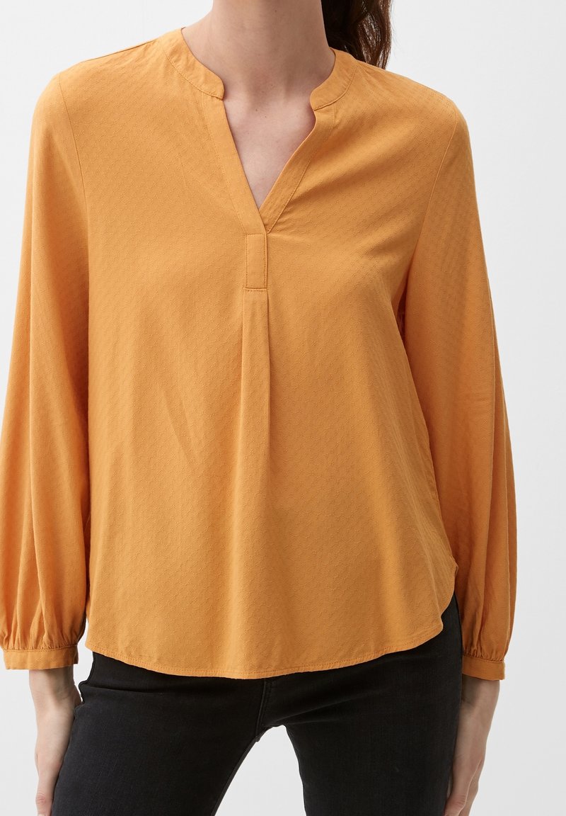 Blouse jaune moutarde avec col en V, détail plissé et manches longues. Tissu doux avec une texture subtile. Portée avec un pantalon noir.