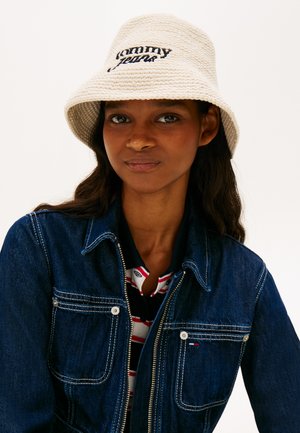 Kobieta w kremowym dzianinowym kapeluszu typu bucket z napisem "Tommy Jeans", ciemnoniebieskiej jeansowej kurtce i czarnej koszulce w paski w kolorach czarnym, białym i czerwonym.