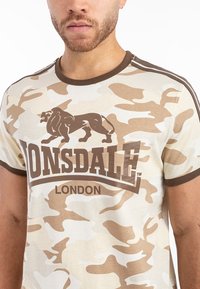 Kreminės spalvos kamufliažo marškinėliai trumpomis rankovėmis, rudi kraštai ir liūto logotipas. Po logotipu užrašyti ryškūs "LONSDALE" ir "LONDON" tekstai.