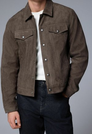 Homme portant une veste en daim marron à boutons sur une chemise blanche et un jean foncé, avec une main dans la poche, sur un fond uni.