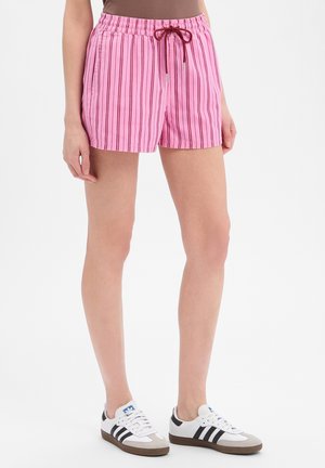 Vrouw draagt roze gestreepte korte broek met een bordeauxroze trekkoord en witte sneakers met zwarte strepen, staand tegen een effen achtergrond.