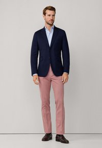 Blazer navy, camicia azzurro chiaro e pantaloni rosa. Il blazer presenta due bottoni e tasche a patta; le scarpe sono in pelle marrone scuro.