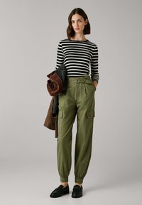 Vrouw in een zwart-wit gestreepte langemouwen top, hoge groene cargobroek, zwarte loafers, met een opgevouwen bruin-zwarte jas in haar hand.