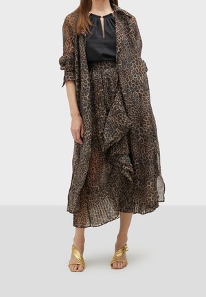 Mujer que lleva una falda larga de leopardo transparente y una chaqueta a juego sobre una camiseta negra con tacones abiertos dorados, de pie sobre un suelo gris.