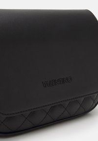 Černá kožená kabelka s prošívaným spodním dílem, zdobená vyraženým logem "Valentino" na přední straně a elegantním zakřiveným klopovým uzávěrem.