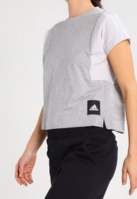 Szara, skrócona koszulka z bawełny, z krótkimi białymi rękawami, bocznymi rozcięciami oraz czarną metką Adidas na dolnym brzegu.