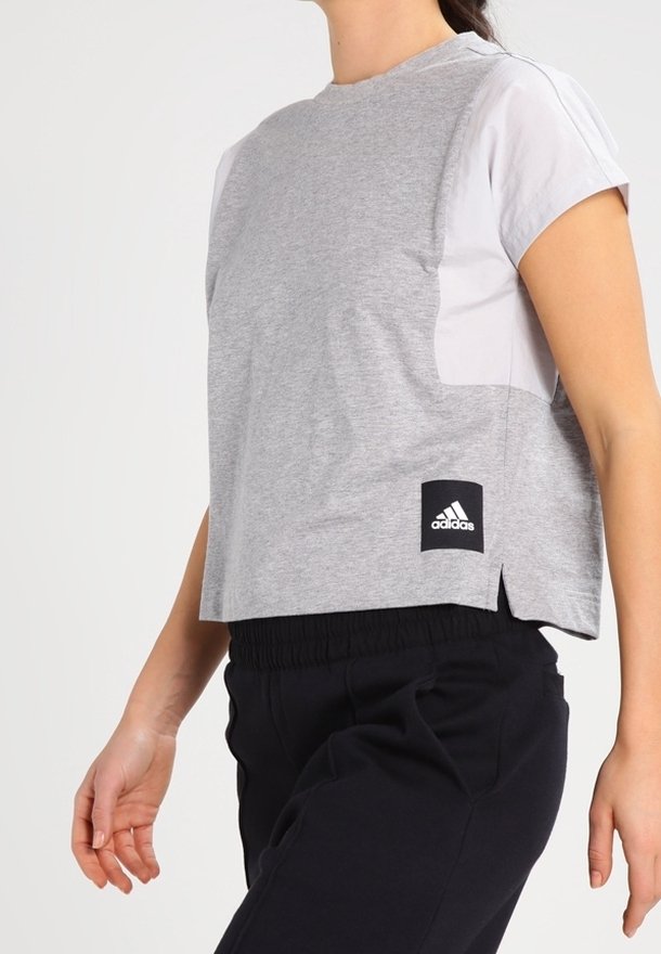 Szara, skrócona koszulka z bawełny, z krótkimi białymi rękawami, bocznymi rozcięciami oraz czarną metką Adidas na dolnym brzegu.