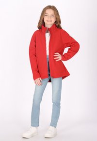Giacca softshell rossa con colletto alto, chiusura a zip e tasche laterali, indossata sopra una camicia bianca e jeans azzurri chiari, abbinata a sneakers bianche.