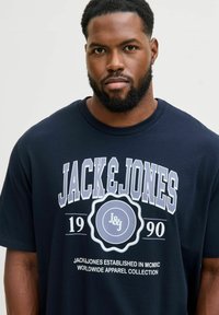 Tumši zils kokvilnas T-krekls ar uzkrītošu balto un gaiši zilo grafiku, uz kuras redzams teksts "JACK & JONES" un dibināšanas gads, ar apļveida kakla dizainu.