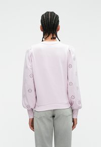 Lila sweatshirt met ballonmouwen en uitsnijdingen in bloemenmotieven. Ribbelkraag en -manchetten. Gecombineerd met lichtgrijze jeans.