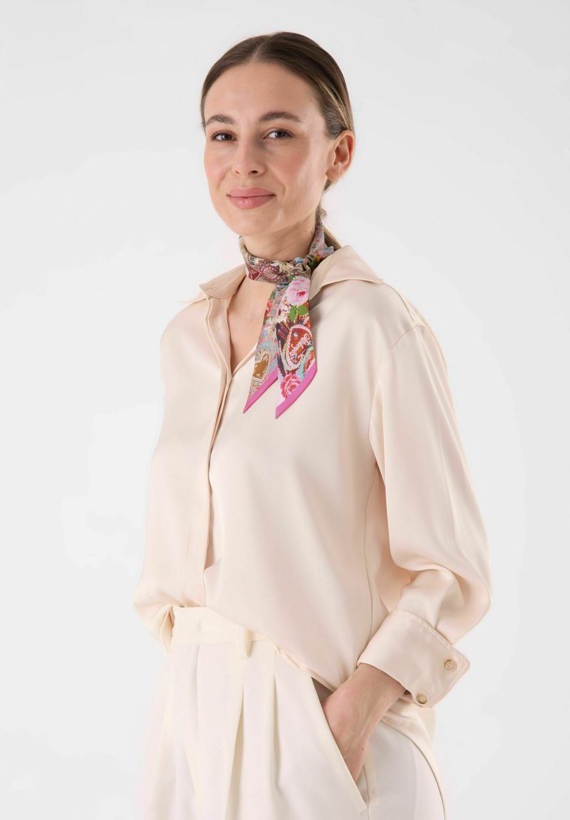 Chemise boutonnière beige clair avec une texture douce. Une écharpe en soie à motif dans des tons de rose et de marron accentue le cou. Les pantalons ont des plis.