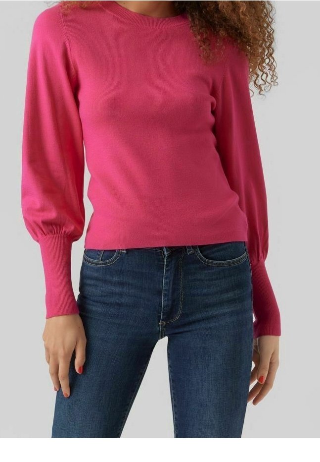 Femme portant un pull moulant rose vif à manches longues bouffantes et un jean bleu foncé sur un fond uni.
