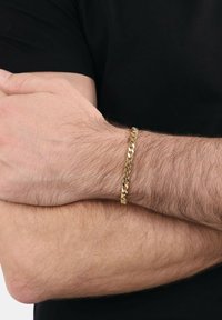 Hombre con los brazos cruzados, vistiendo una camiseta negra y una pulsera de eslabones dorados en la muñeca izquierda.