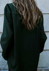 Dark green oversized shirt with long sleeves, featuring a smooth texture and button cuffs, seen from the back against a stone wall.