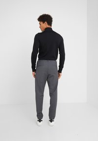 Schwarzer Pullover mit langen Ärmeln und hohem Kragen, kombiniert mit grauen, maßgeschneiderten Hosen und weißen Sneakers. Der Stoff wirkt glatt und figurbetont.
