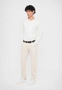 Fehér, hosszú ujjú, V-nyakú póló, bézs nadrág, fekete öv és fehér sportcipő. A modell a zsebébe tett kézzel áll egy sima háttér előtt.