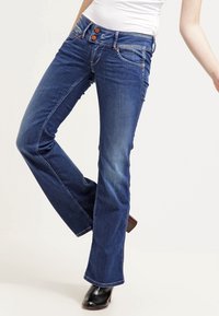 Kvinna som bär midrise mörkblå bootcut-jeans med orange knappar, i kombination med svarta klackboots och en vit topp.