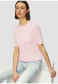 Roze gebreide top met korte mouwen en een ronde halslijn, met een gladde textuur en een subtiele logo-detail aan de zoom, gecombineerd met lichtblauwe jeans.