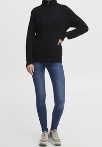 Schwarzer Strickpullover mit hohem Kragen und Reißverschluss, kombiniert mit blauen engen Jeans und grauen Turnschuhen. Glatte Textur und lässiges Design.