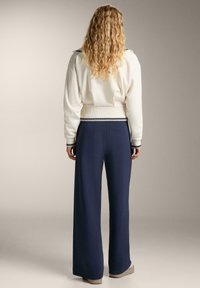 Pulover alb din tricot cu tiv ondulat, purtat împreună cu pantaloni largi bleumarin. Ambele au accente contrastante și o siluetă lejeră.