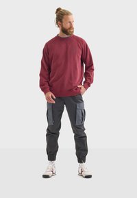 Felpa bordeaux con collo rotondo, abbinata a pantaloni cargo neri con accenti in grigio, e sneakers bianche con dettagli rossi.