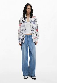 Weißes Hemd mit Knöpfen und mehrfarbigem grafischem Text, getragen über einem weißen Tanktop und blauen Jeans mit weitem Bein. Schwarze und weiße Sneakers vervollständigen den Look.