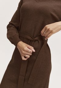 Abito a maglia marrone con scollatura a V e maniche lunghe, dotato di una cintura di tessuto legata in vita. Tessuto liscio e vestibilità comoda con polsini a costine.
