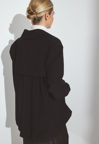 Femme avec des cheveux blonds en chignon portant un manteau noir ample et un col roulé blanc, vue de dos sur un fond clair.