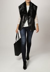 Schwarze Lederweste mit einem Kunstfellkragen, kombiniert mit einem hellen Pullover, dunklen Jeans und schwarzen ankle boots. Schwarze Tote-Tasche in der Hand.