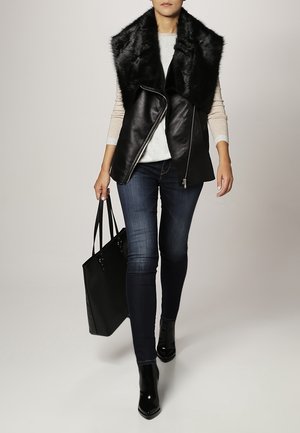 Gilet en cuir noir avec un col en fausse fourrure, associé à un pull léger, un jean sombre et des bottines noires. Sac fourre-tout noir à la main.