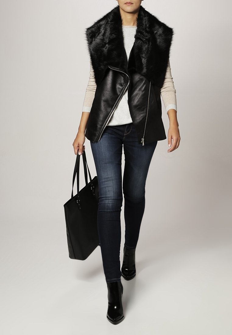 Schwarze Lederweste mit einem Kunstfellkragen, kombiniert mit einem hellen Pullover, dunklen Jeans und schwarzen ankle boots. Schwarze Tote-Tasche in der Hand.