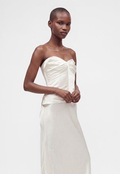 Femme portant une robe bustier en satin blanc avec un détail froncé sur le buste, debout devant un fond clair uni.