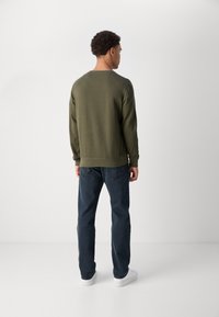 Olejnatě zelená mikina s kulatým výstřihem a žebrovanými manžetami, v kombinaci s tmavě modrými džíny a bílými teniskami. Jednoduchý design, bez vzorů.