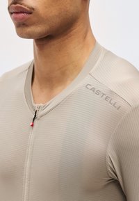 Κοντινό πλάνο σε άτομο που φοράει μπεζ, υφασμάτινο πουκάμισο με φερμουάρ και το λογότυπο "CASTELLI" στον δεξιό ώμο.